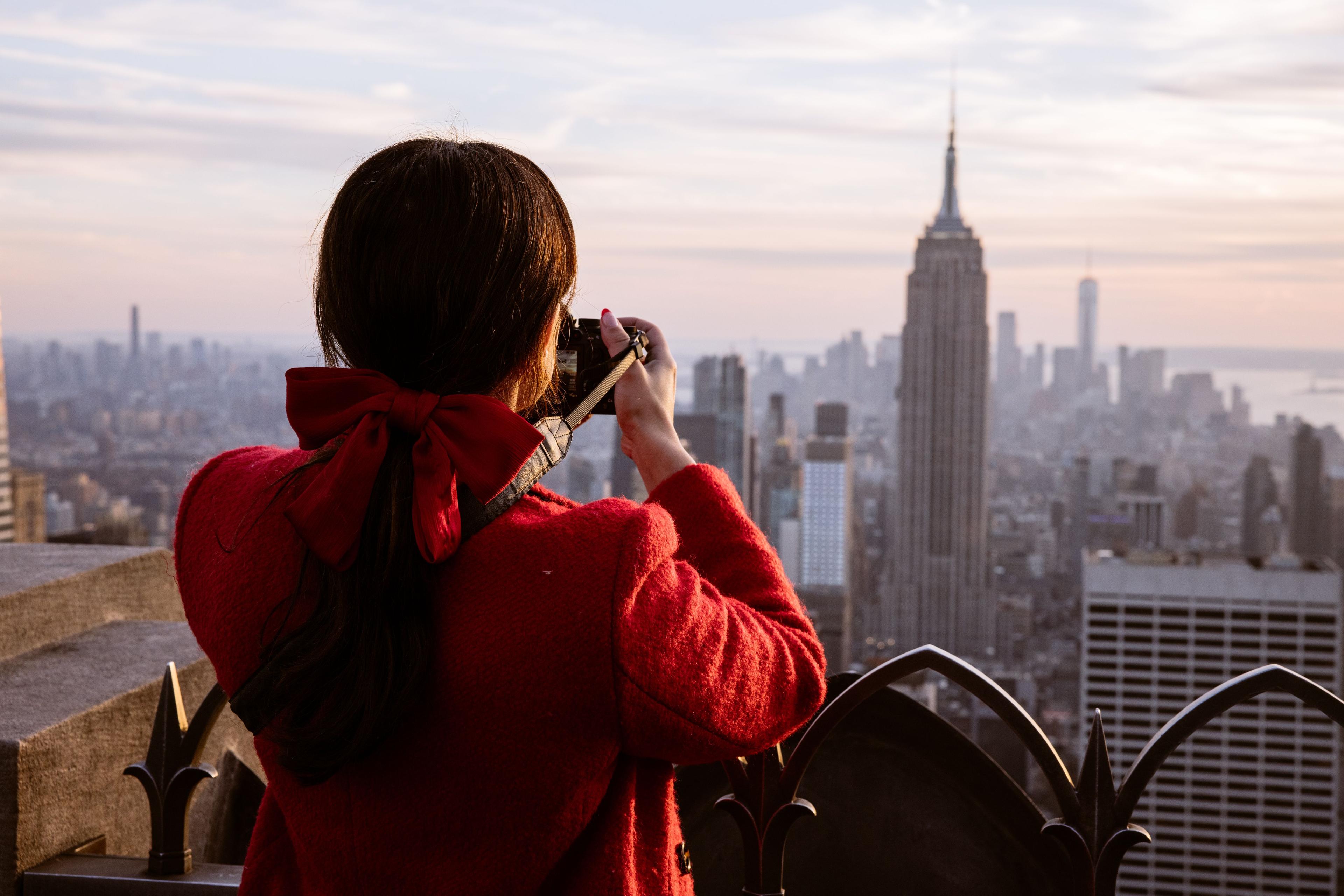 a person with a red bow in their ponytail taking a photo of NYC from the top of the rock. They are wearing a red sweater