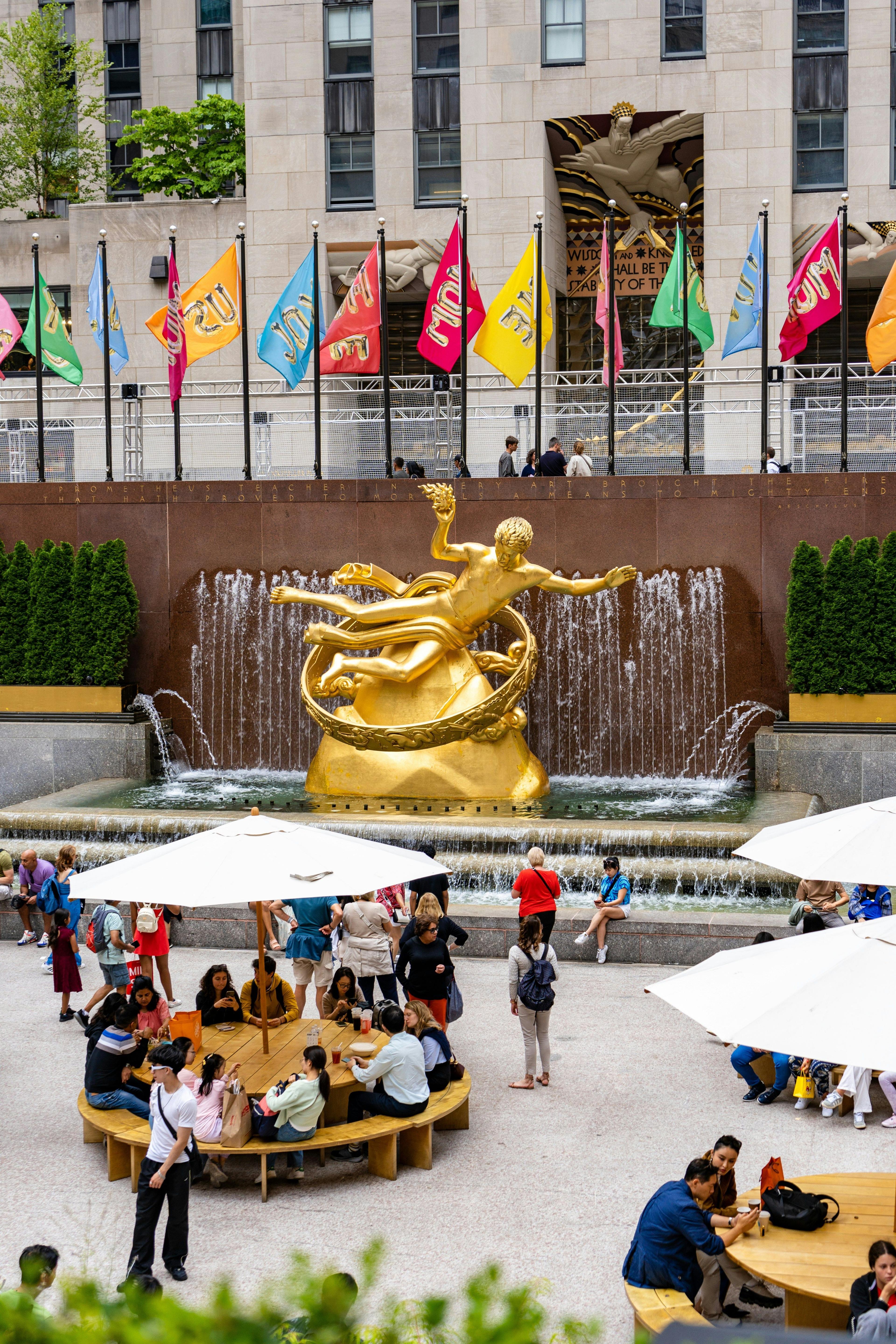 The Prometheus statue and fountain at Rockefeller Center, with people at outdoor tables under umbrellas.