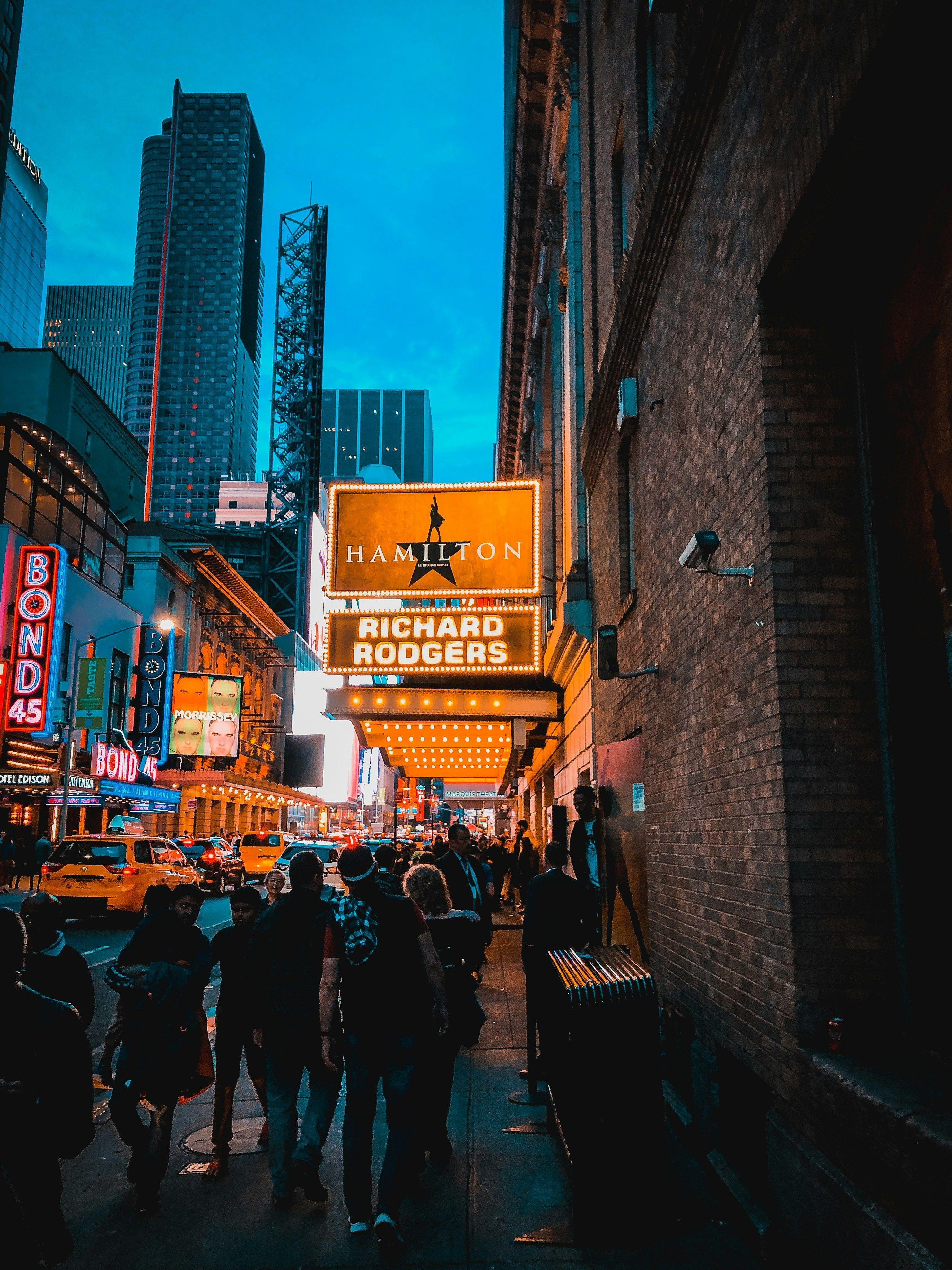 A lit Broadway theater marquee at dusk, with people walking below.